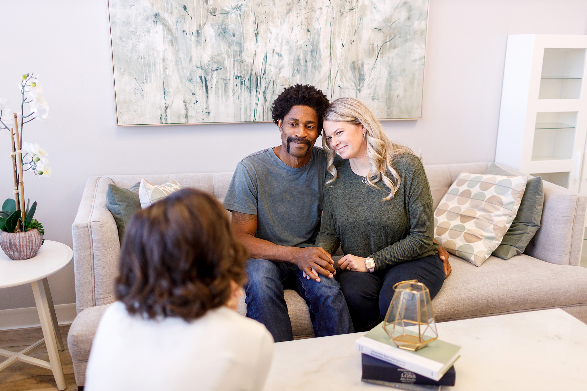 A couple smiles as they hold hands in a side-by-side embrace. They are sitting on a couch in a couples therapy session.