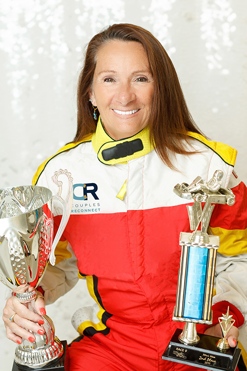 A woman wearing a race car suit smiles as she holds up two gold trophies,