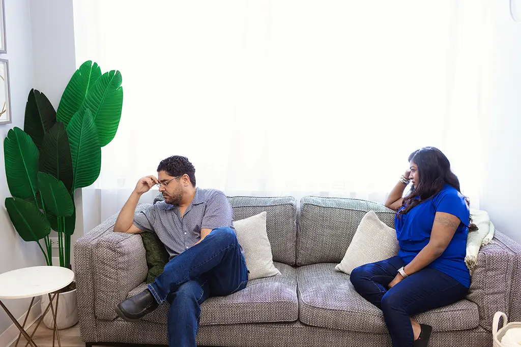 A man and a woman sitting on the left and right side of a couples therapy couch. The man is looking away, to the left, while holding his hand to his face. The woman is looking in his direction.