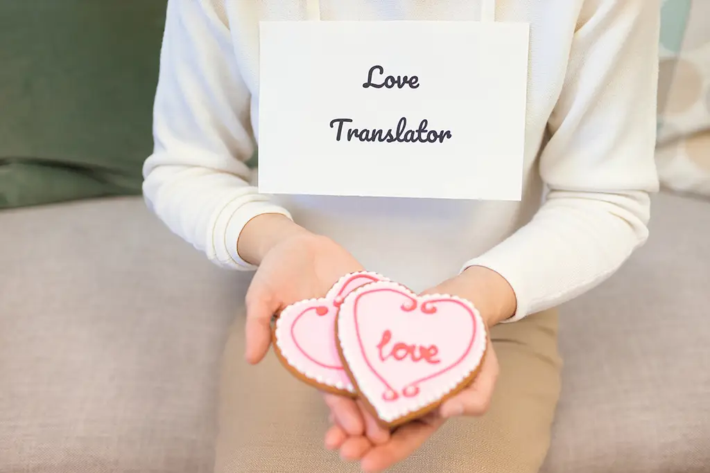 A woman holding two cookies decorated with icing that say "Love". Around her neck, she is wearing a sign that says "Love Translator".