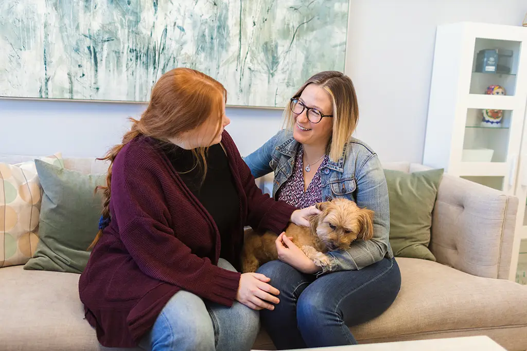 Two women sitting on a couples therapy couch and holding a small dog.