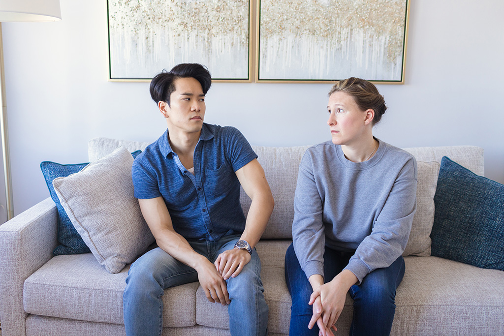 A man and a woman frown as they look at each other. They are sitting side by side on a therapy couch.