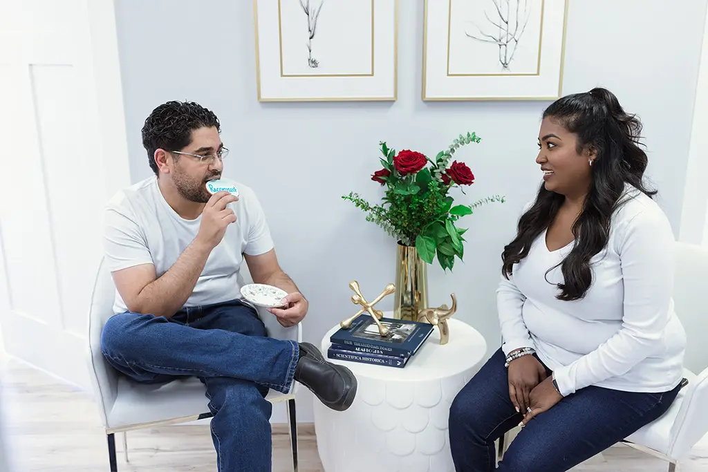 A man and a woman are seated facing each other. He is eating a cookie, while she talks. Between them there is a vase with a bouquet of red roses.