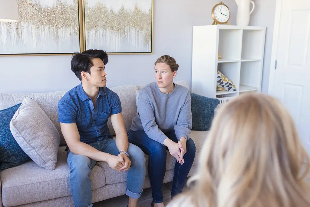 A man and a woman sitting side by side in a couples therapist office. The expressions on their faces reflect emotional distress. The back of the therapist's head appears in the foreground.