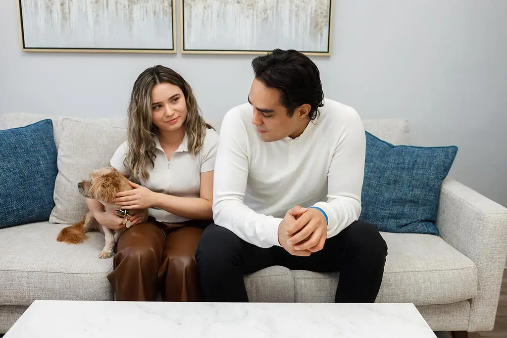 A woman and a man looking at each other, as they sit side by side on a couples counselling couch. The woman, on the left, is holding a small dog in her lap.