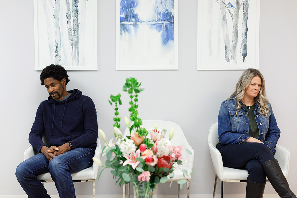 A couple sit in a waiting room, facing away from each other. The man is sitting on the left chair, looking down and facing left. There is an empty chair in the middle. On the right, a woman is sitting on a chair, looking down and to the right.