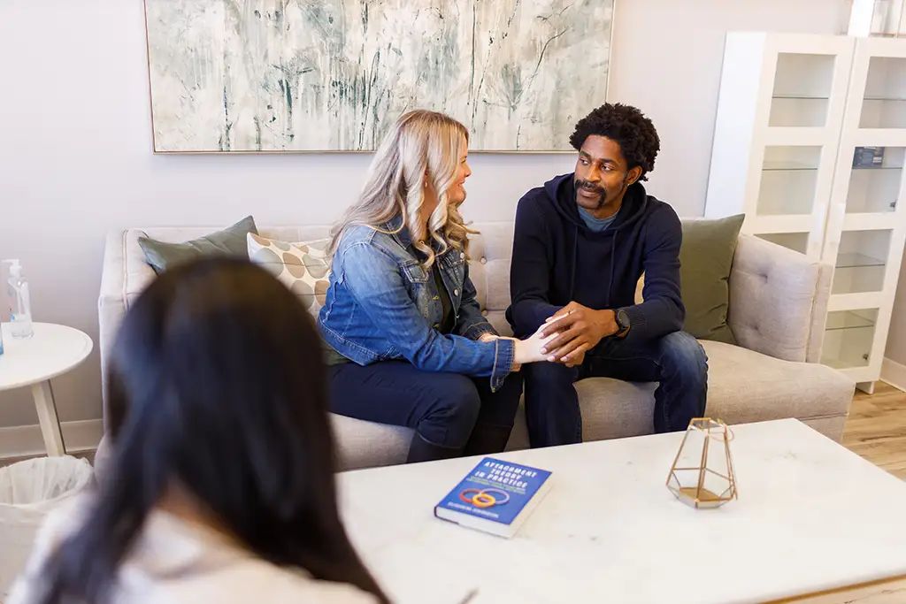 A woman and a man sitting on a couples counselling couch. The back of a therapist's head is visible in the bottom left corner, in the foregraound.