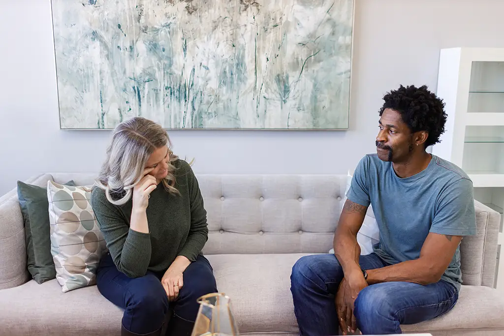 A couple sit side by side on a couples counselling couch. The woman is crying, while her partner looks at her from a distance.