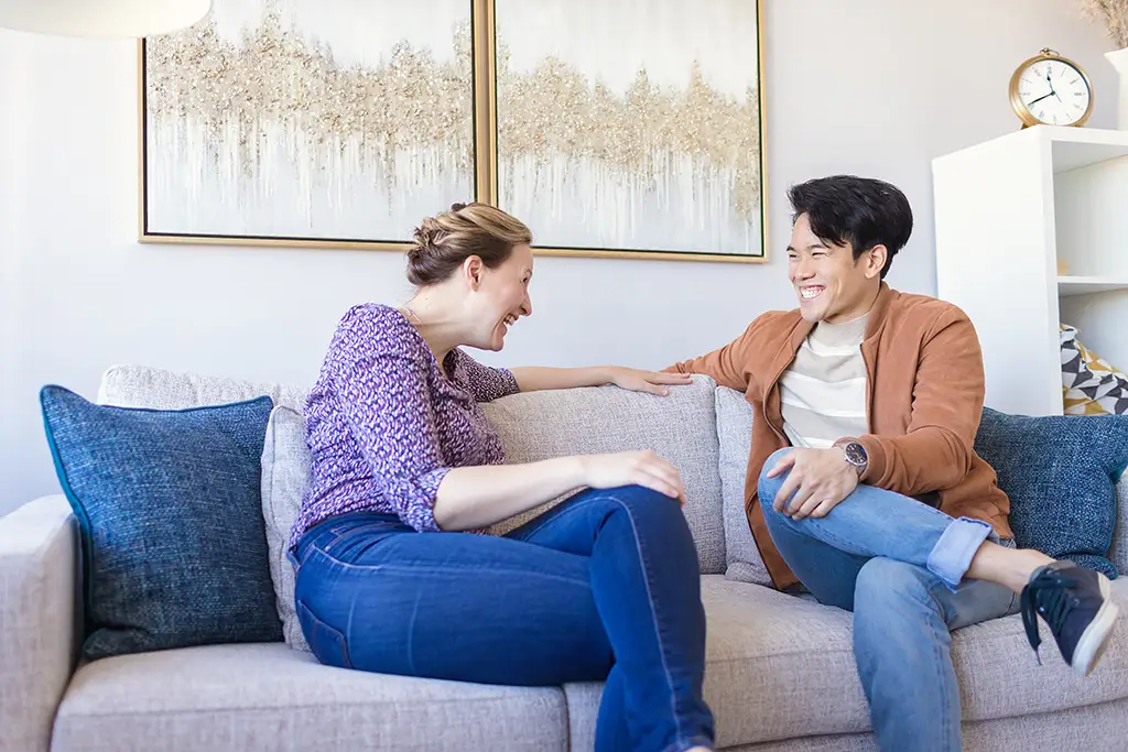 A woman and a man sitting side by side on a couch laugh together during a couples counselling session.