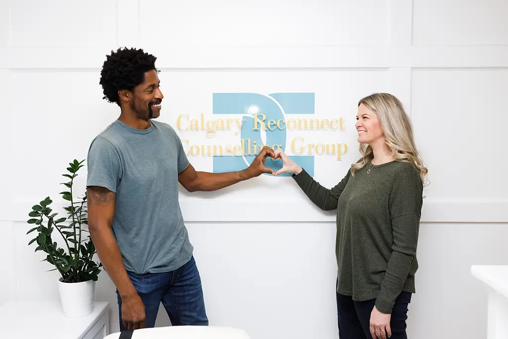 A man and a woman stand in front of a sign on the wall that says "Calgary Reconnect Counselling Group”. They are looking at each other. He is holding out his left hand and she is holding up her right hand; together, they are forming a heart shape.