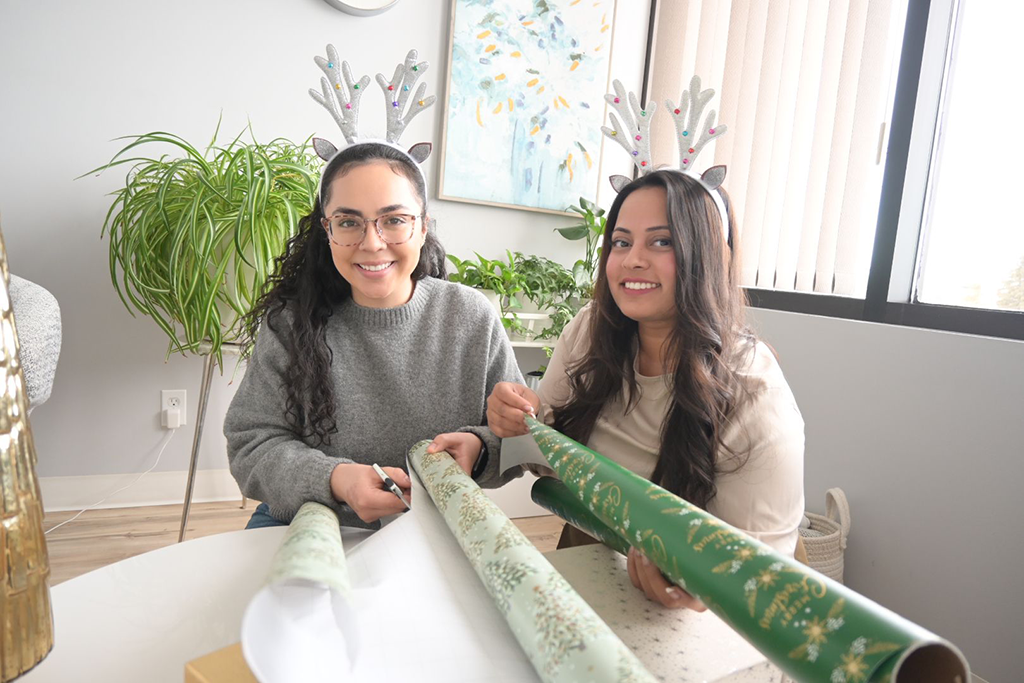 Two women wearing reindeer antlers smile at the camera as they wrap Holiday presents.