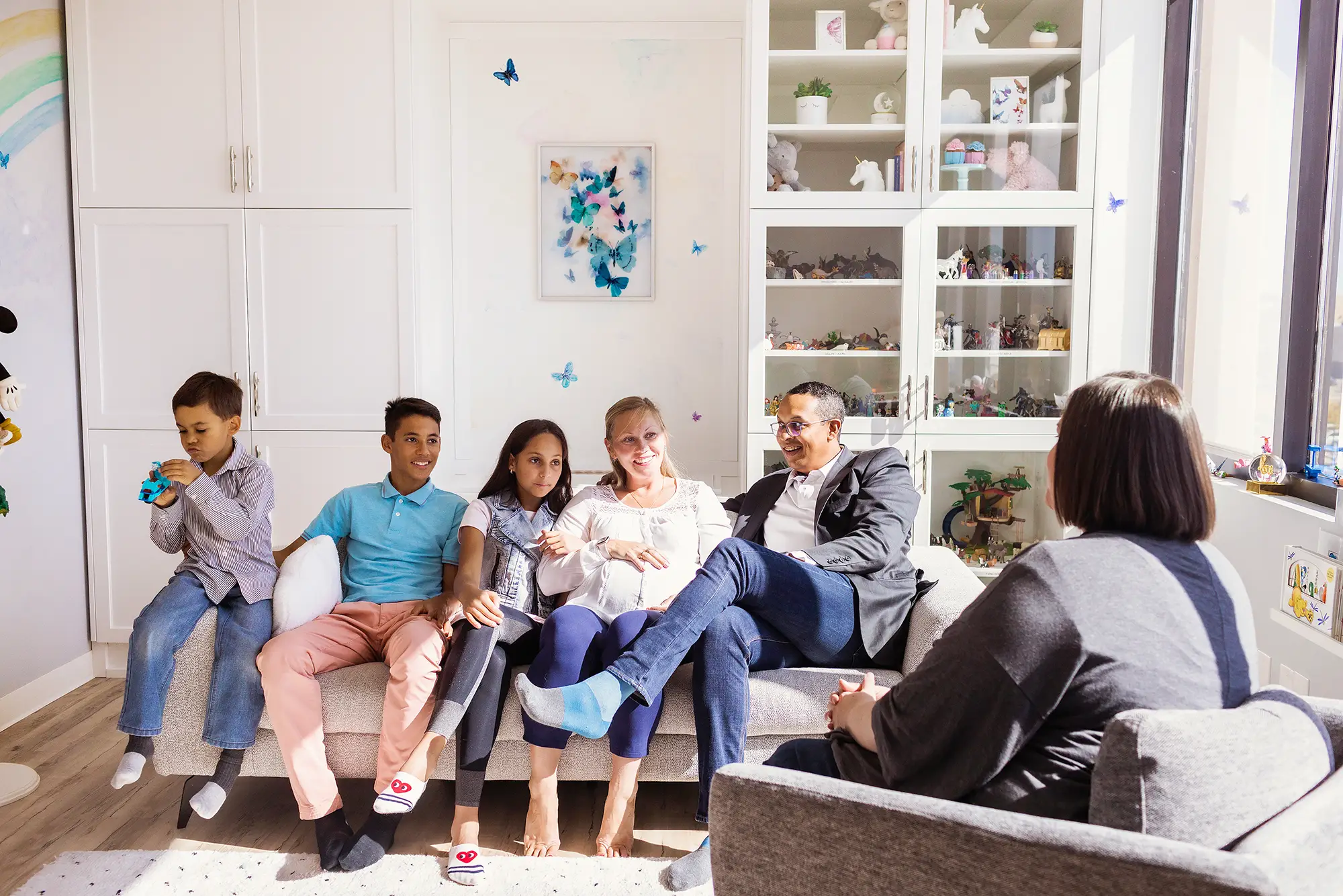 We have everyone covered A family counselling session in a playroom at Kids Reconnect. From left to right: two boys, a girl, a mother, and a father sitting on a therapy couch in a playroom. To the right, a family therapist in the foreground.