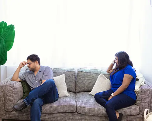 A man and a woman are sitting far apart on a couples therapy couch. On the left, the man is holding on hand against his forehead, facing away from the woman. The woman is sitting on the right, looking at him.