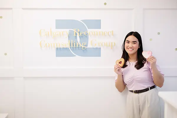 Couples Reconnect is a branch of Calgary Reconnect Counselling Group A woman smiles as she holds a heart-shaped cookie in each hand. She is standing in front of a wall sign that reads: "Calgary Reconnect Counselling Group"