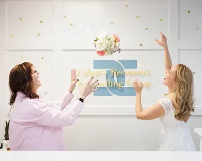 A woman on the left holds her hands in the air as she gestures to catch a bridal bouquet in mid-air, which the woman on the right, couples therapist Natalie Bergman, is tossing. They are standing in front of a sign that reads