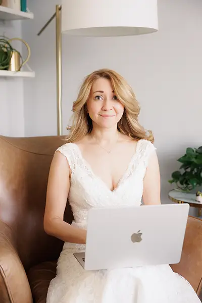 A woman (couples therapist Natalie Bergman), wearing a bridal gown, is sitting on a chair and smiling as she holds a laptop on her lap.