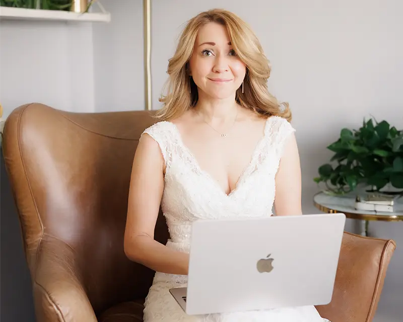 Wedding season is coming – Couples Reconnect A woman (couples therapist Natalie Bergman), wearing a bridal gown, is sitting on a chair and smiling as she holds a laptop on her lap.